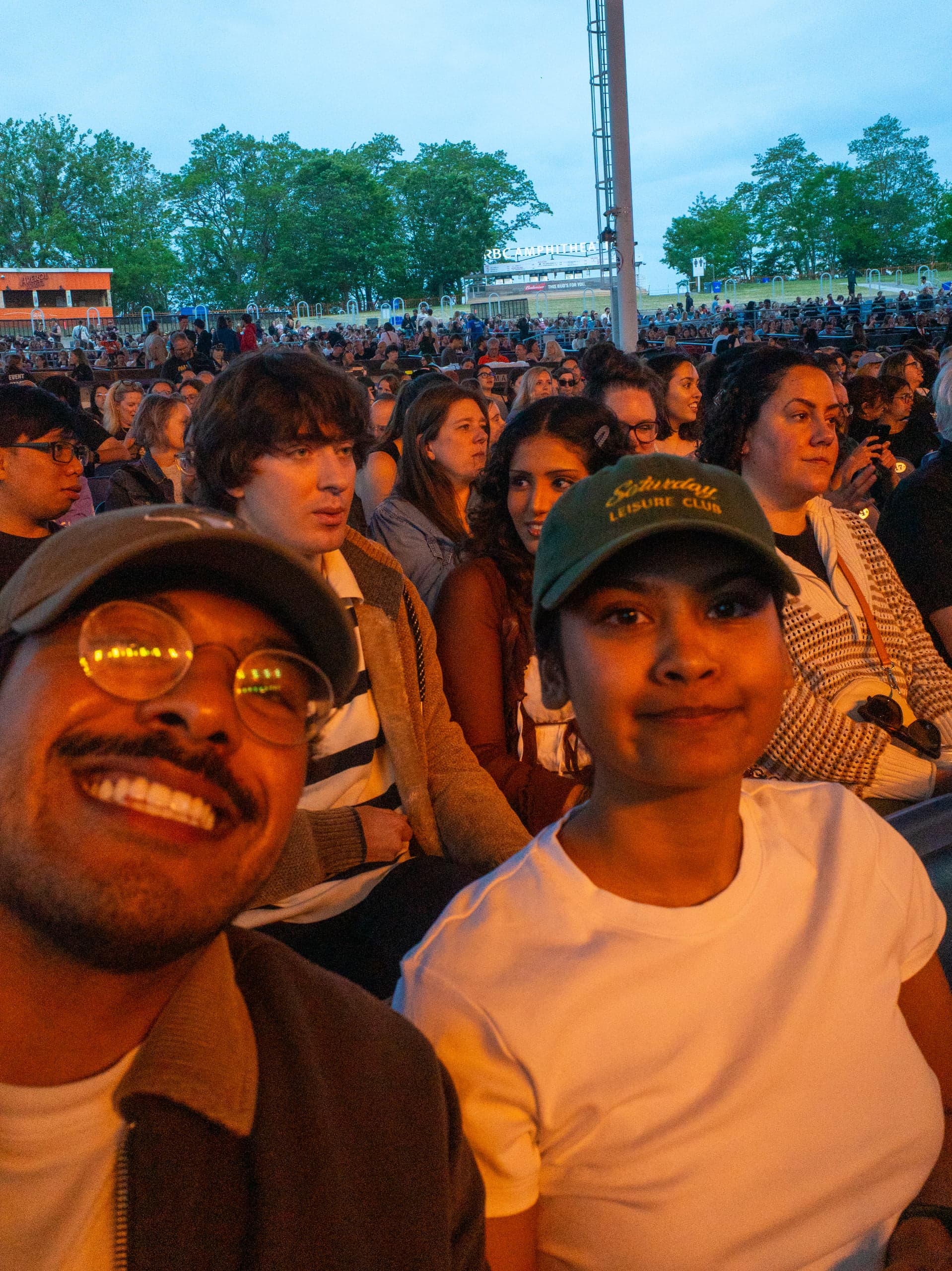 Selfie of two people in a packed outdoor amphitheatre crowd at dusk, before the stage lights are up