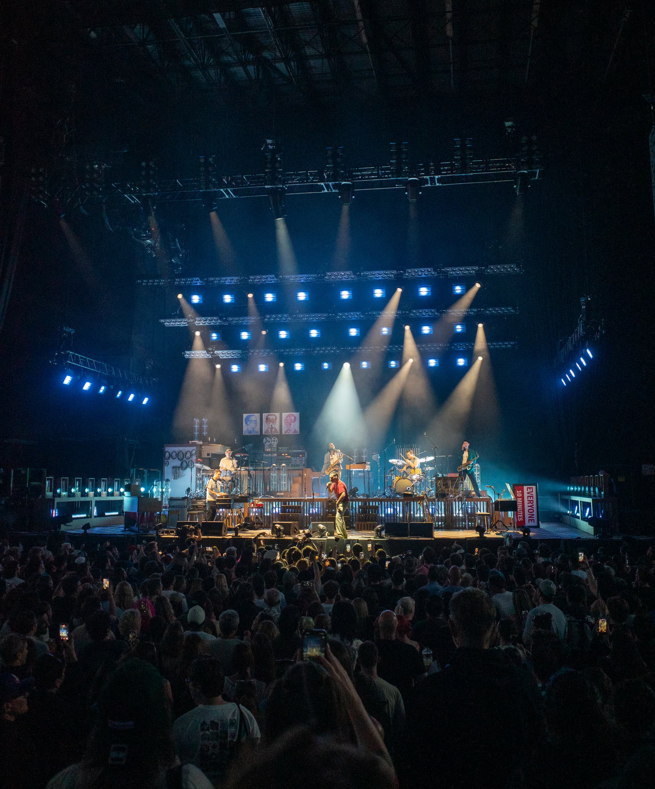 Concert stage under a grid of blue spotlights, the band mid-set, crowd silhouettes with phones raised