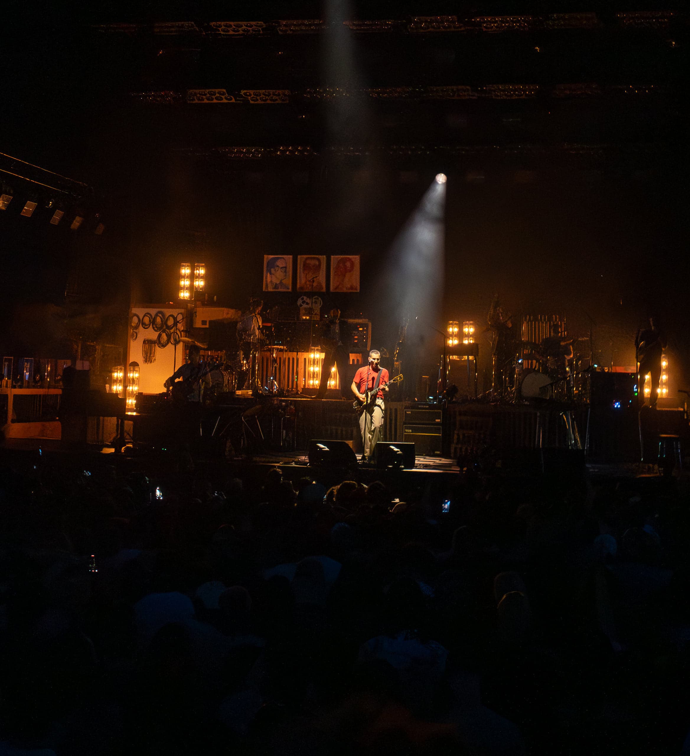 A lone musician at a center mic under a single white spotlight beam, dark crowd in the foreground
