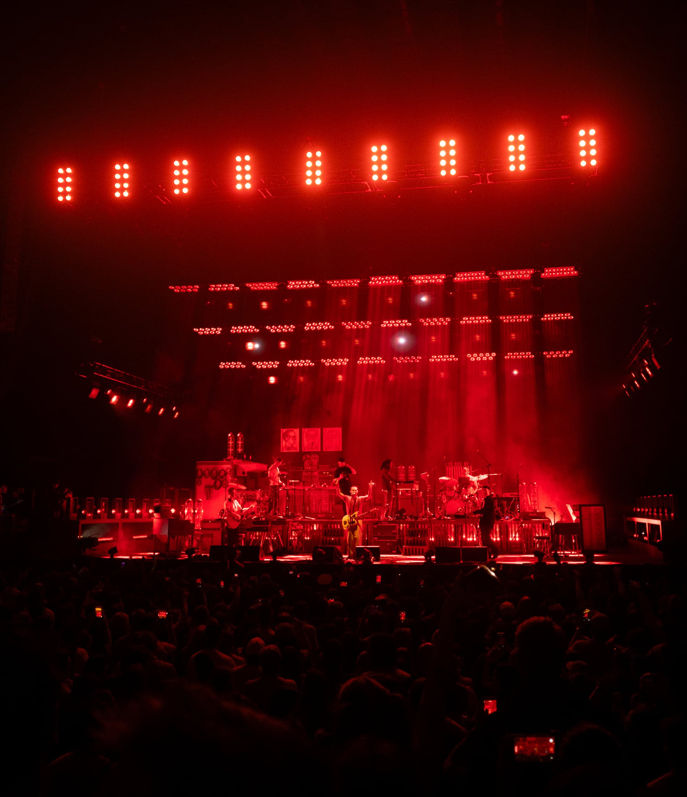 A full stage drenched in deep red light beneath a row of red lamps, band playing to a silhouetted crowd