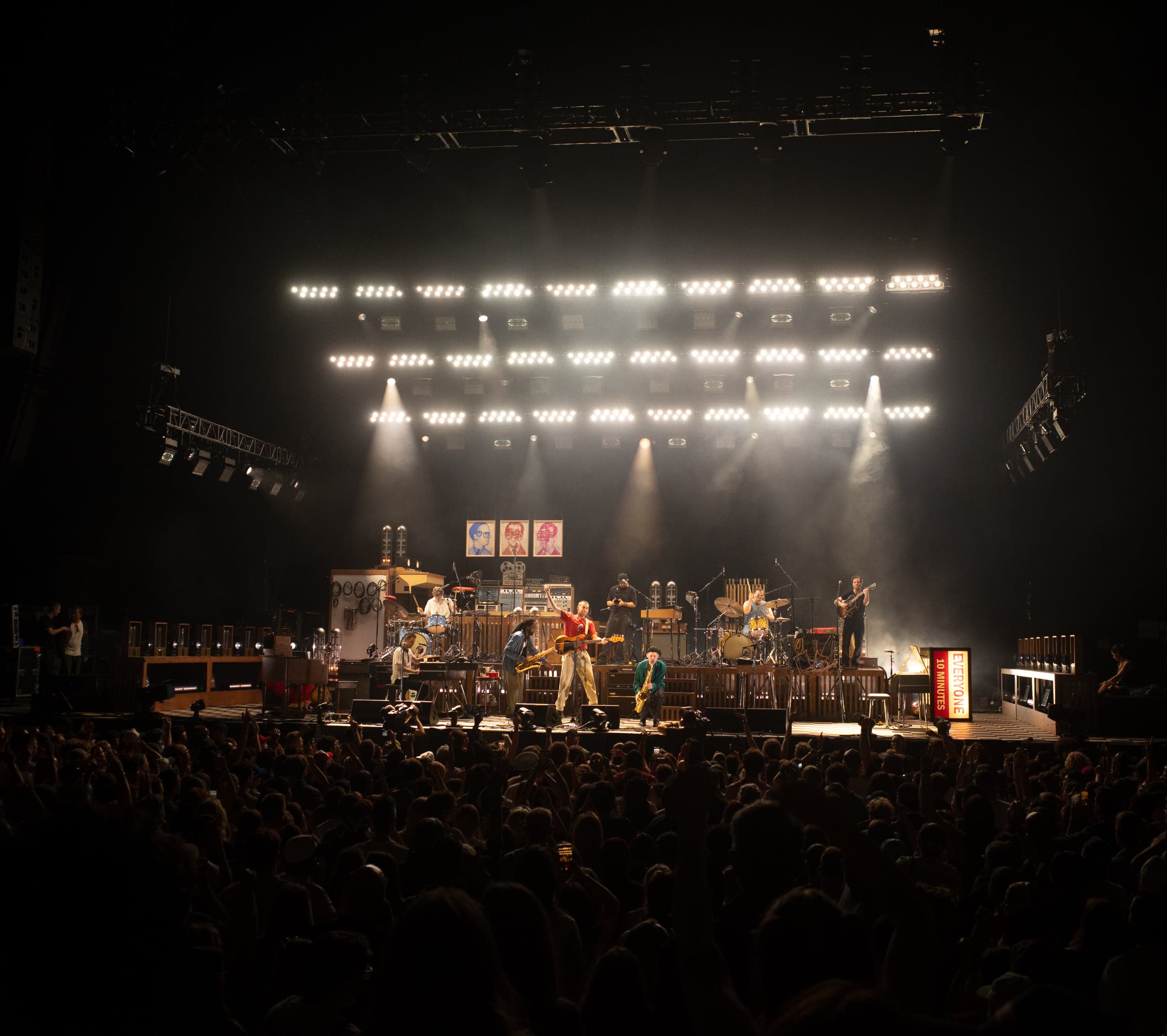 The full band spread across a brightly lit stage under banks of white light, seen over the crowd