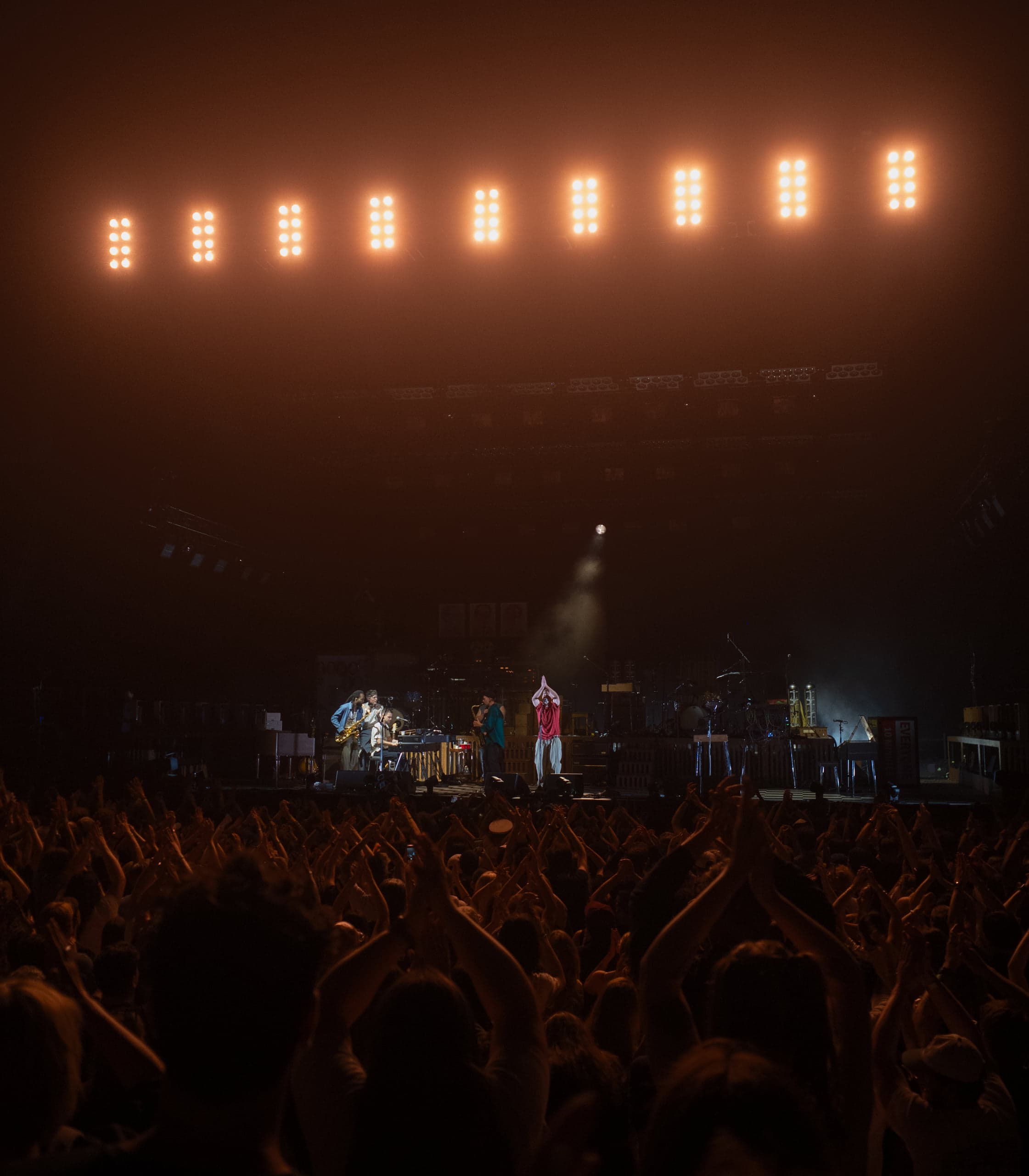 Hands raised across a dark crowd toward a distant hazy stage lit by a row of warm orange lamps