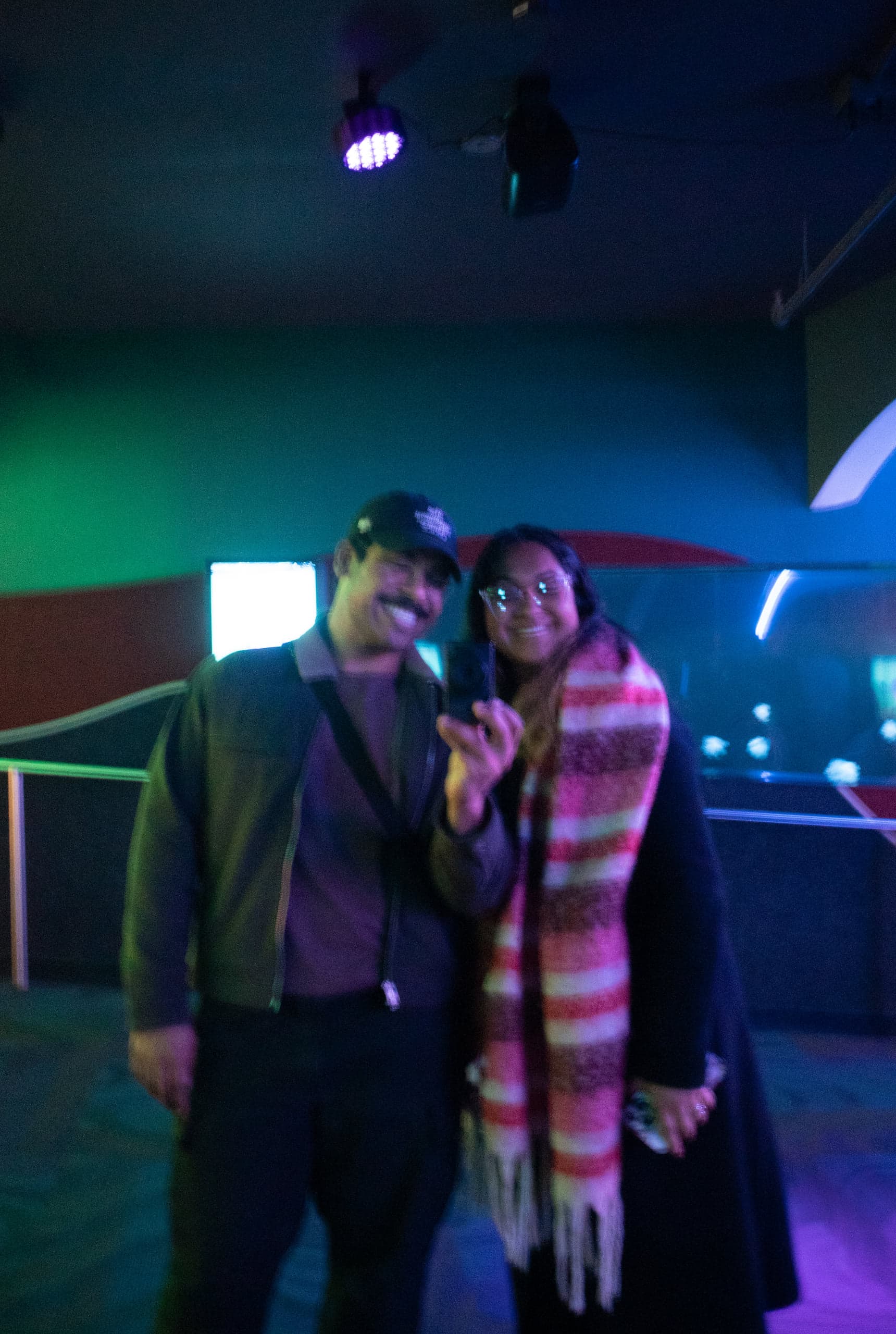 A smiling man and woman posing together in a dark exhibit hall lit green and blue, a tank behind
