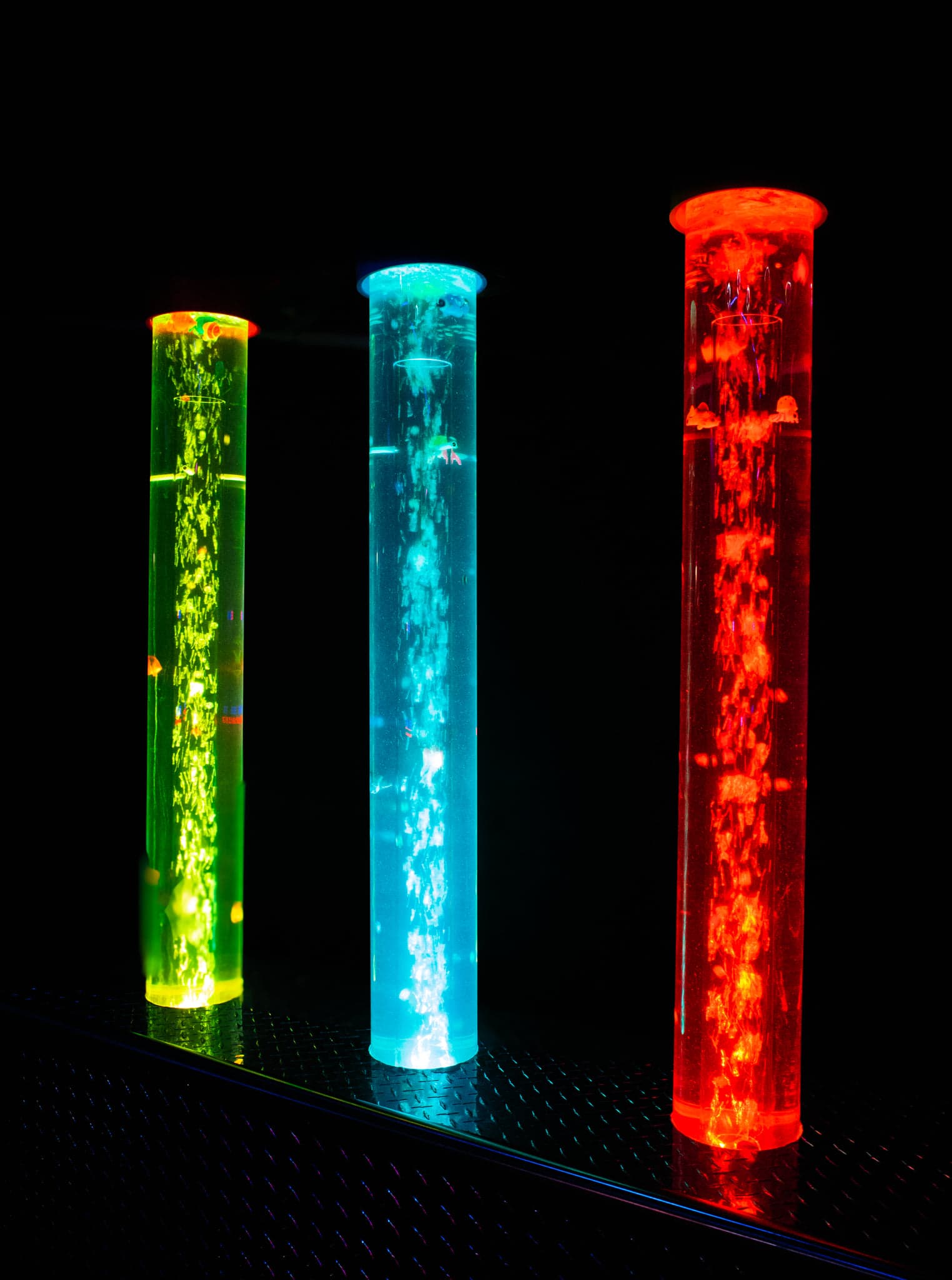 Three tall illuminated tubes glowing green, blue, and red on a dark metal surface against black