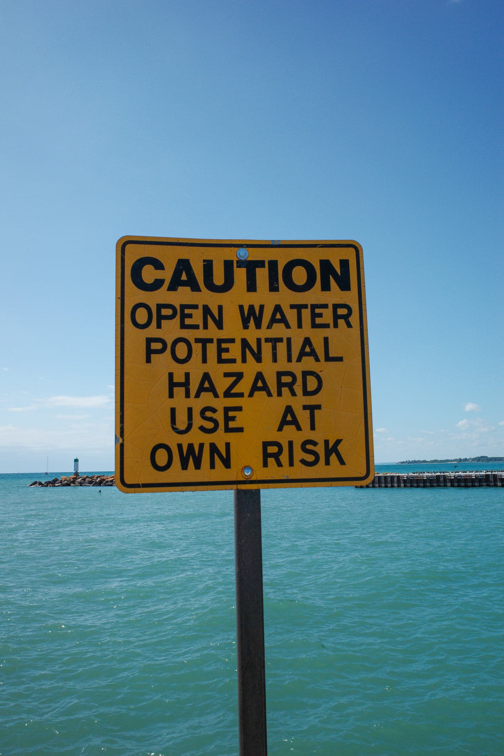 A weathered yellow sign reading CAUTION OPEN WATER POTENTIAL HAZARD USE AT OWN RISK on a post above turquoise water, a rocky breakwater and small beacon behind