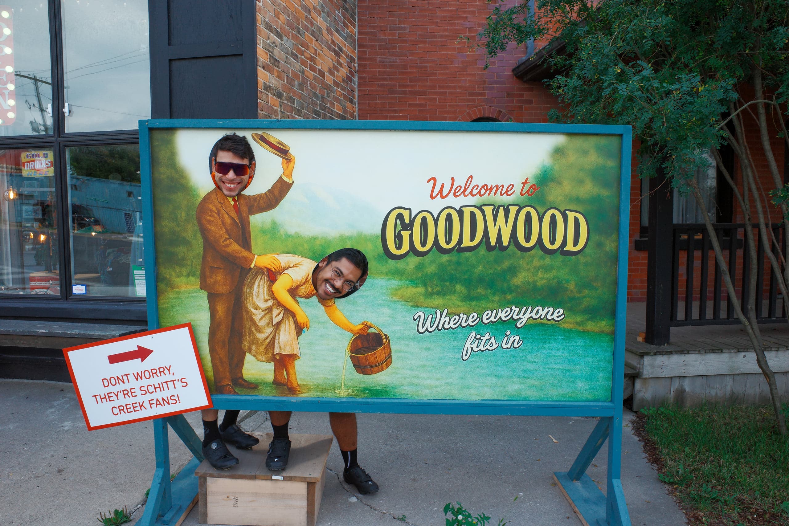 Two people grinning through the face cutouts of a painted board reading 'Welcome to Goodwood, where everyone fits in,' outside a brick storefront