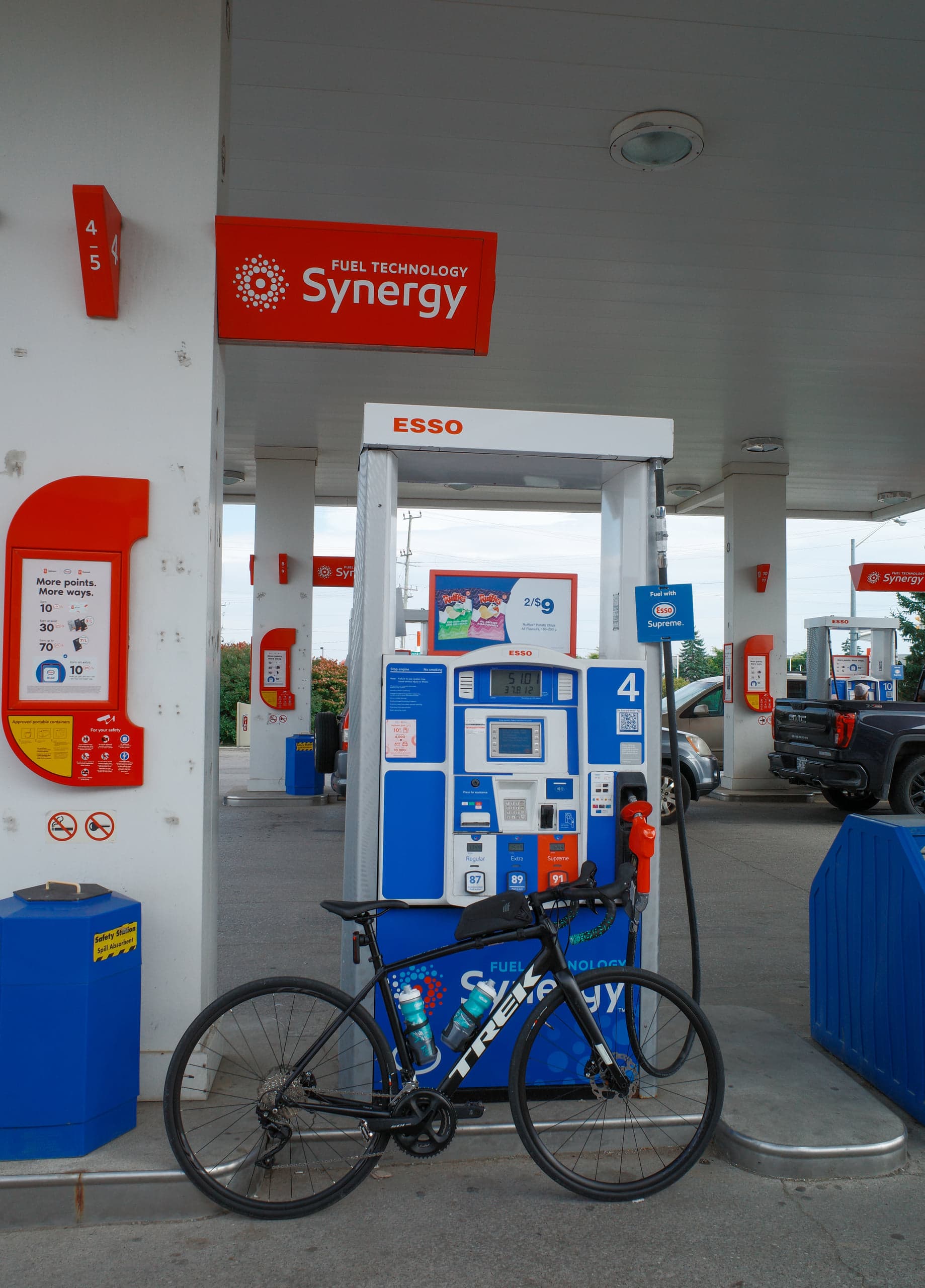 A black Trek road bike with two water bottles leaning against an Esso gas pump under the station canopy, a pickup truck at the pumps beyond