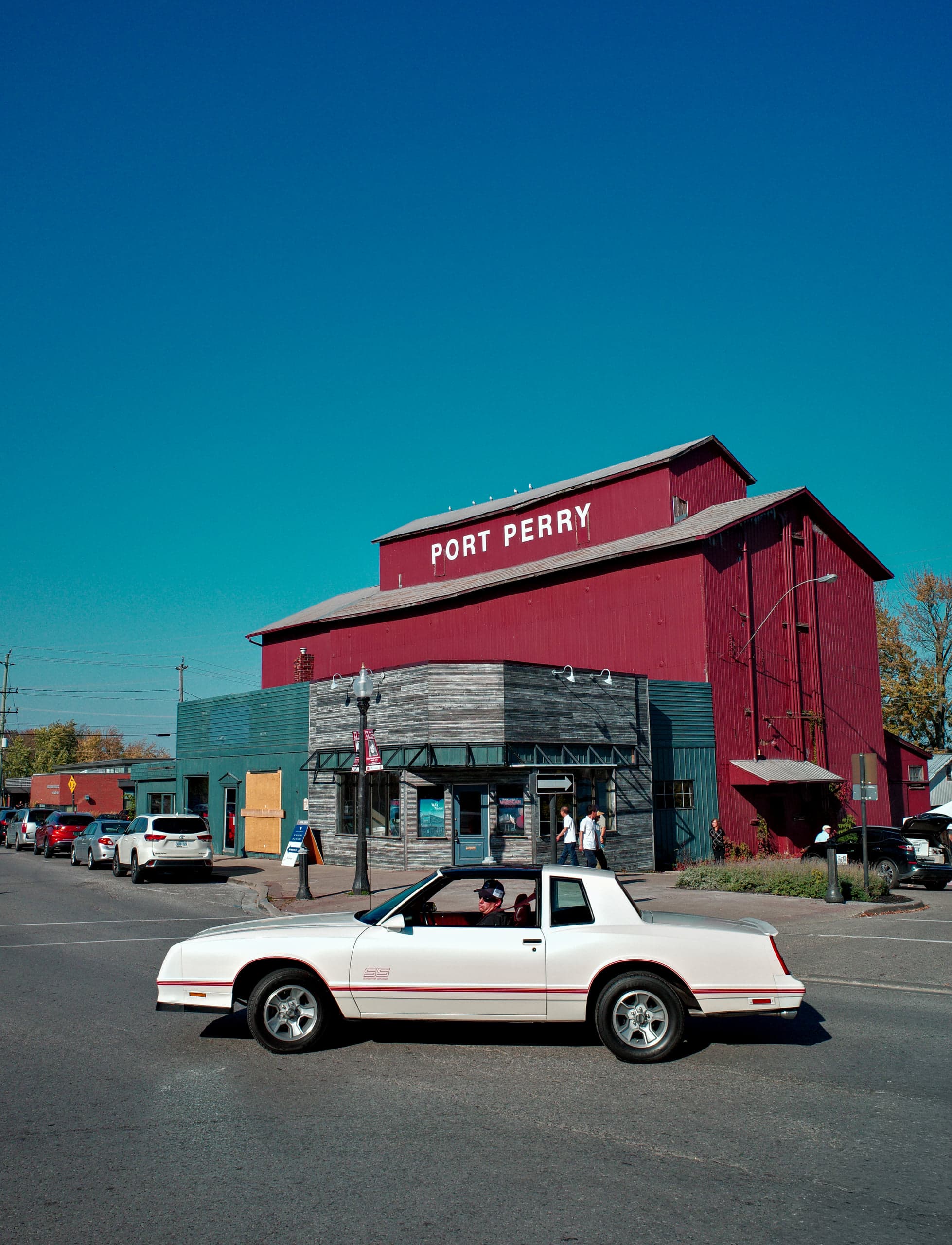 A white 1980s coupe crossing an empty street past the tall red Port Perry feed mill under a clear blue sky