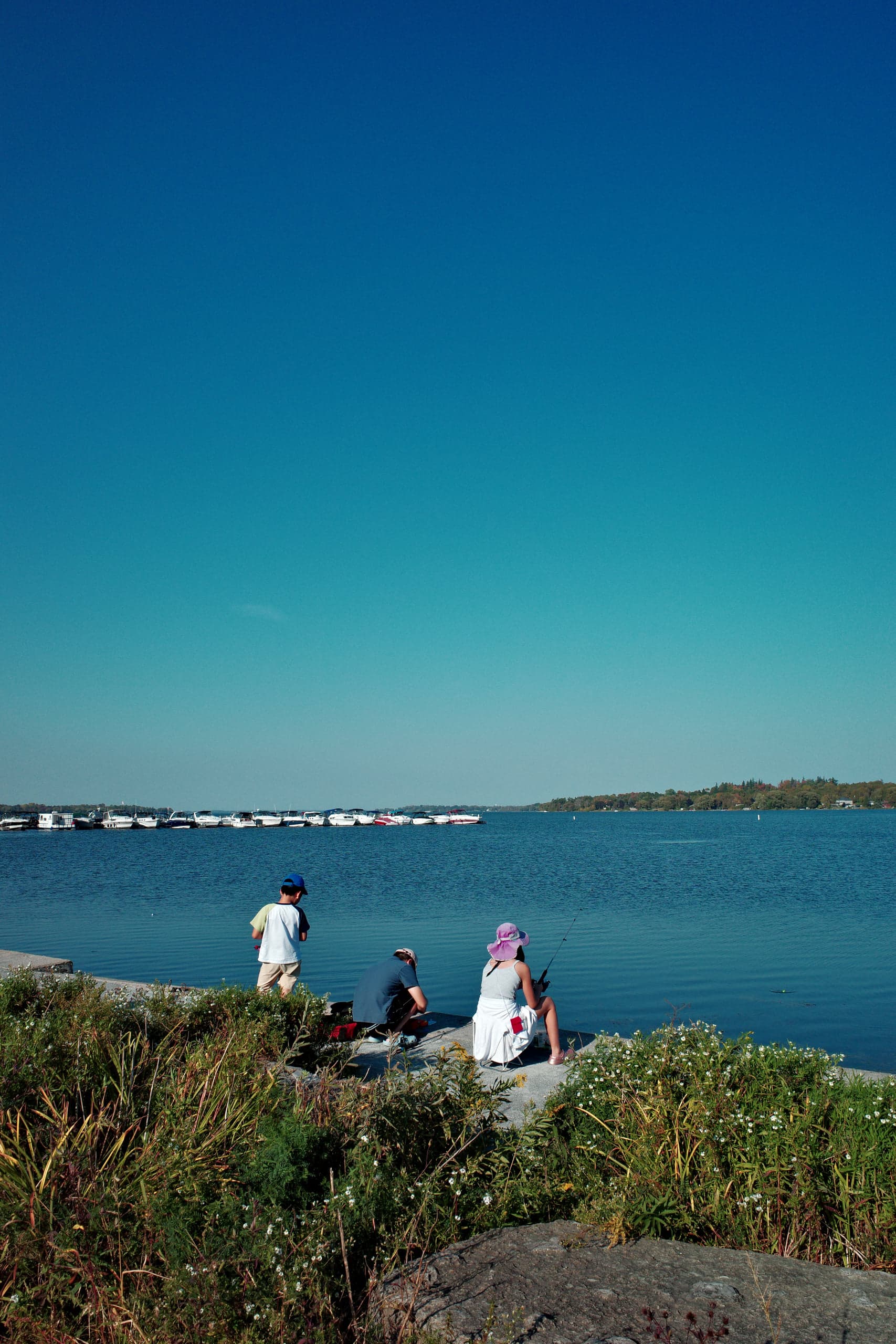 A boy, a crouching adult, and a woman in a sun hat fishing from a low concrete ledge at the water's edge, a marina full of moored boats across the calm lake