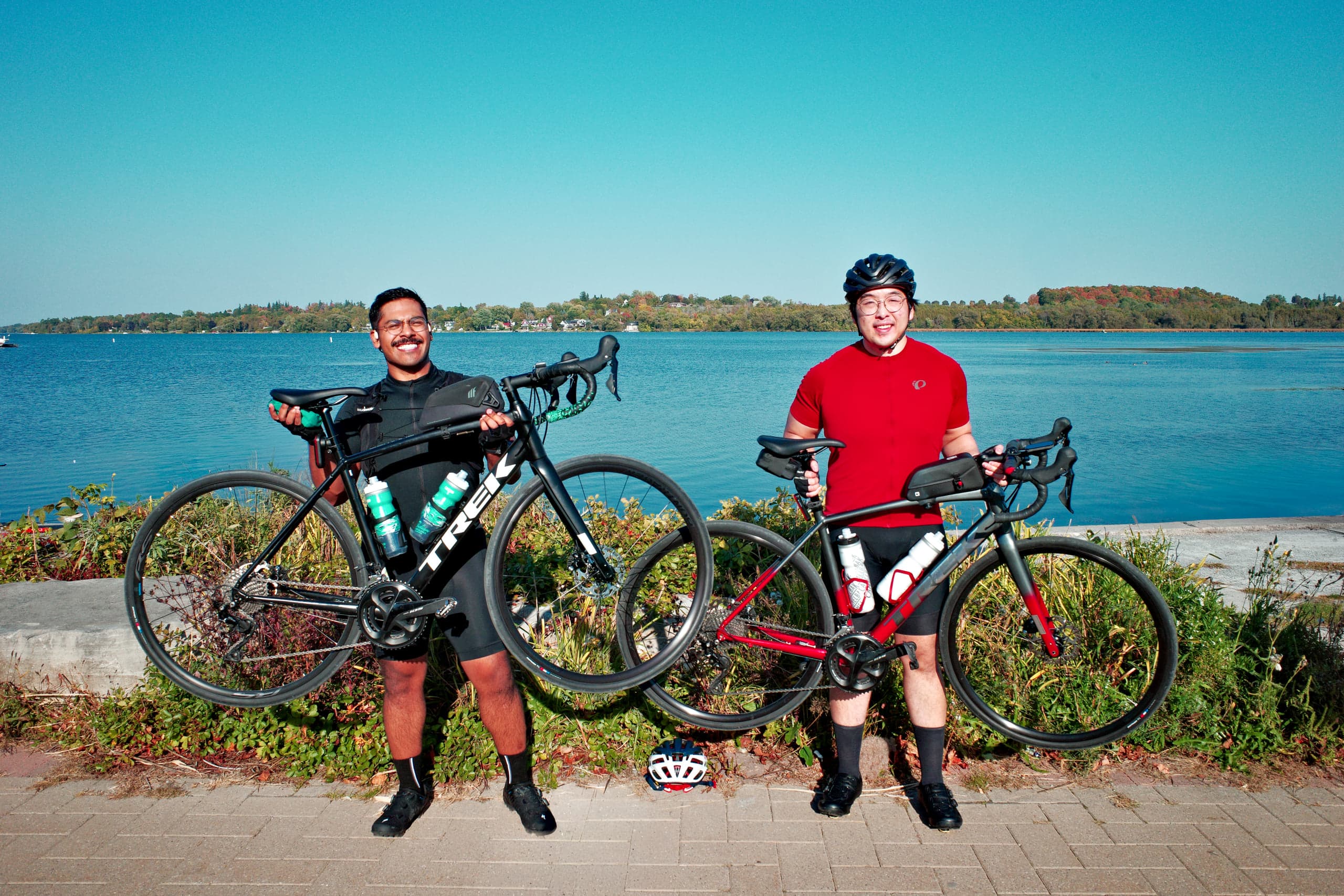 Two cyclists standing on a lakeside path, each lifting a Trek road bike beside them, calm blue water and a tree-lined far shore behind under a clear sky