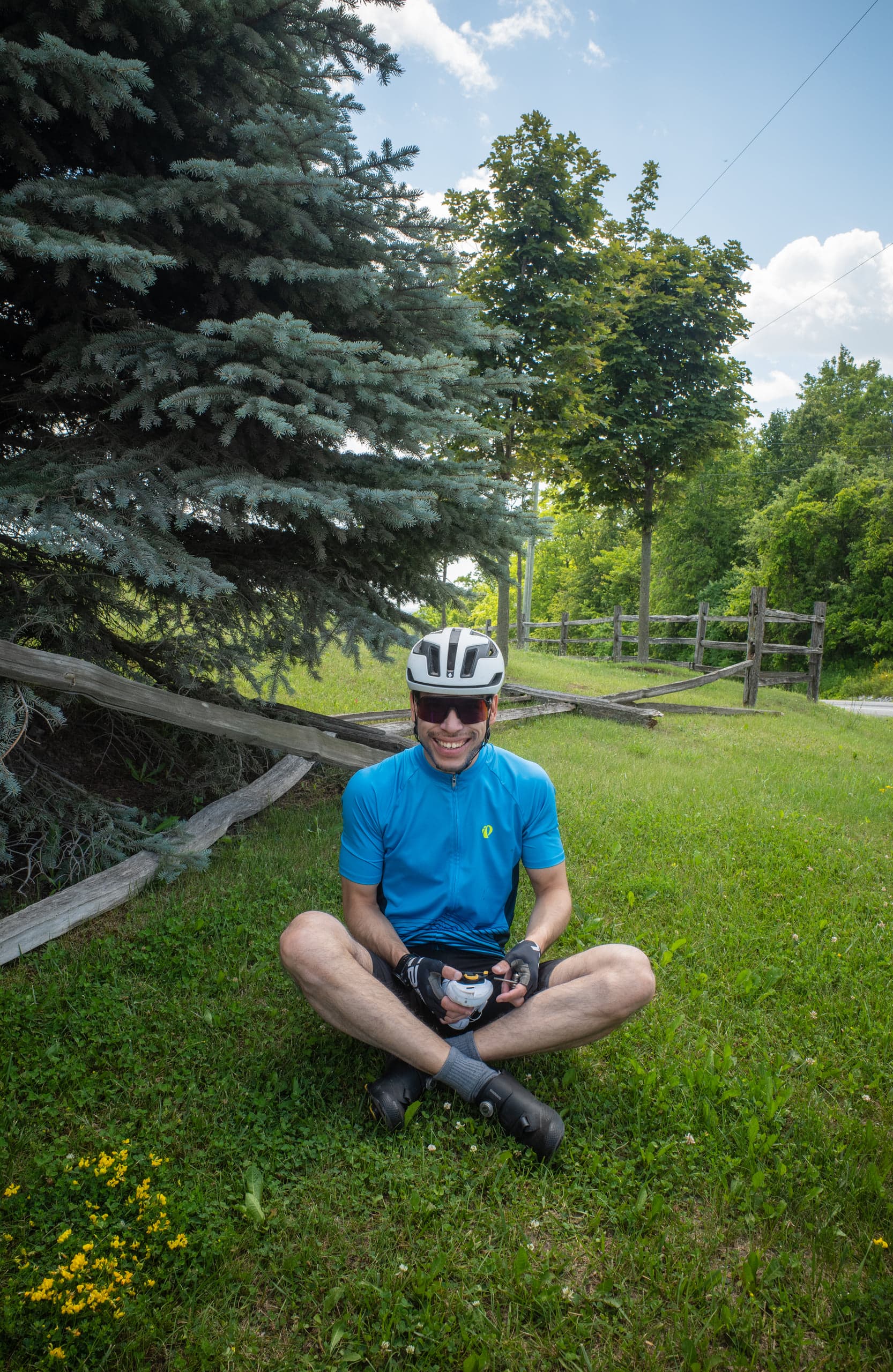 A cyclist in a blue jersey and white helmet sitting cross-legged and smiling on the grass beside a weathered split-rail fence, a large blue spruce and a country road behind