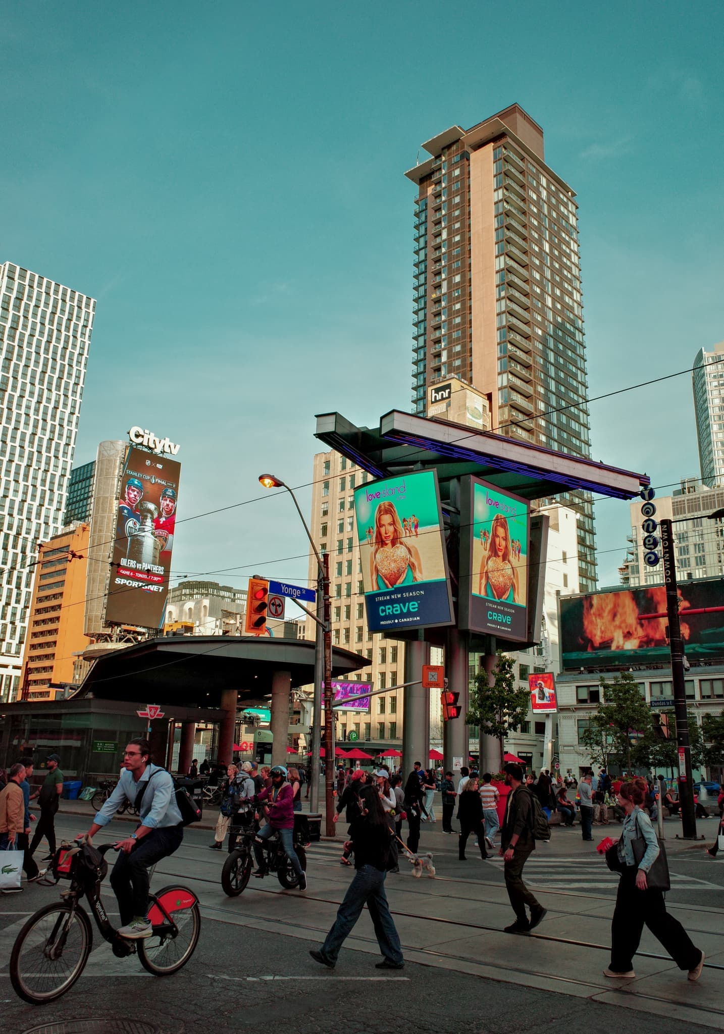 Cyclists and pedestrians cross a downtown intersection past a streetcar shelter topped with glowing billboards at dusk