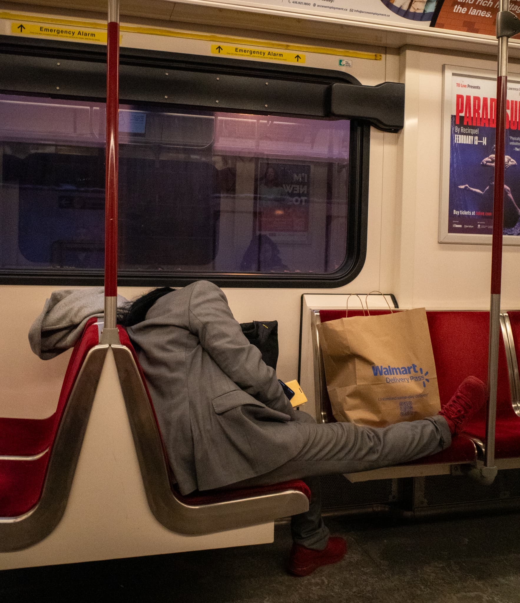 A person sleeps curled across red subway seats beside a Walmart shopping bag, dark window behind them