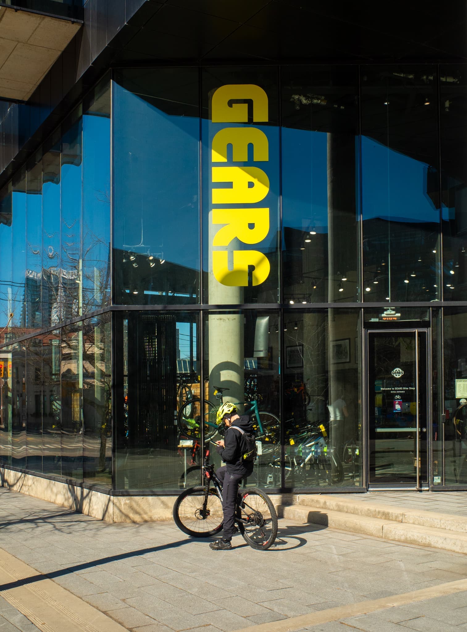A cyclist in a yellow helmet stands with their bike before a glass storefront marked GEARS in yellow letters