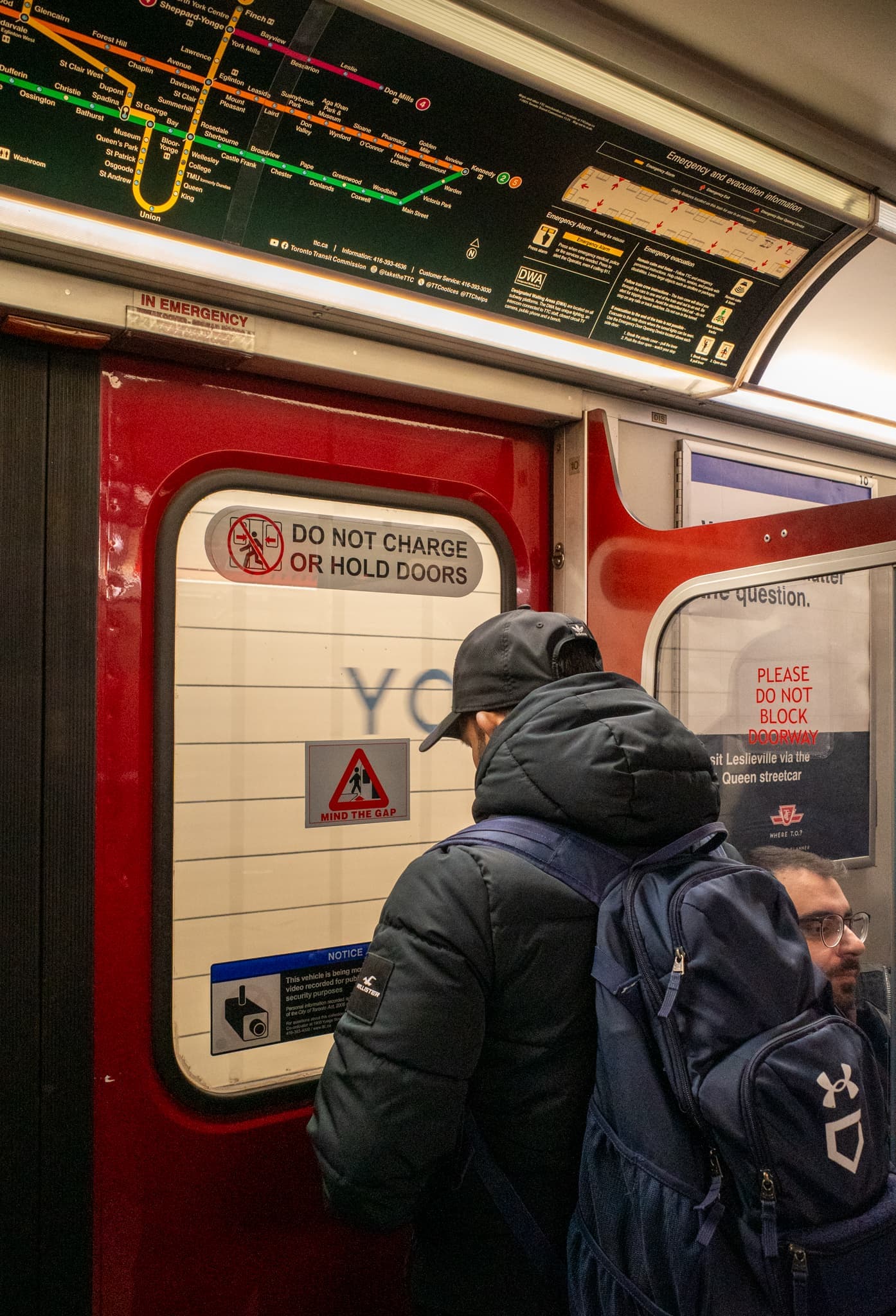 Two commuters in dark winter coats and backpacks wait by red subway doors below a transit route map