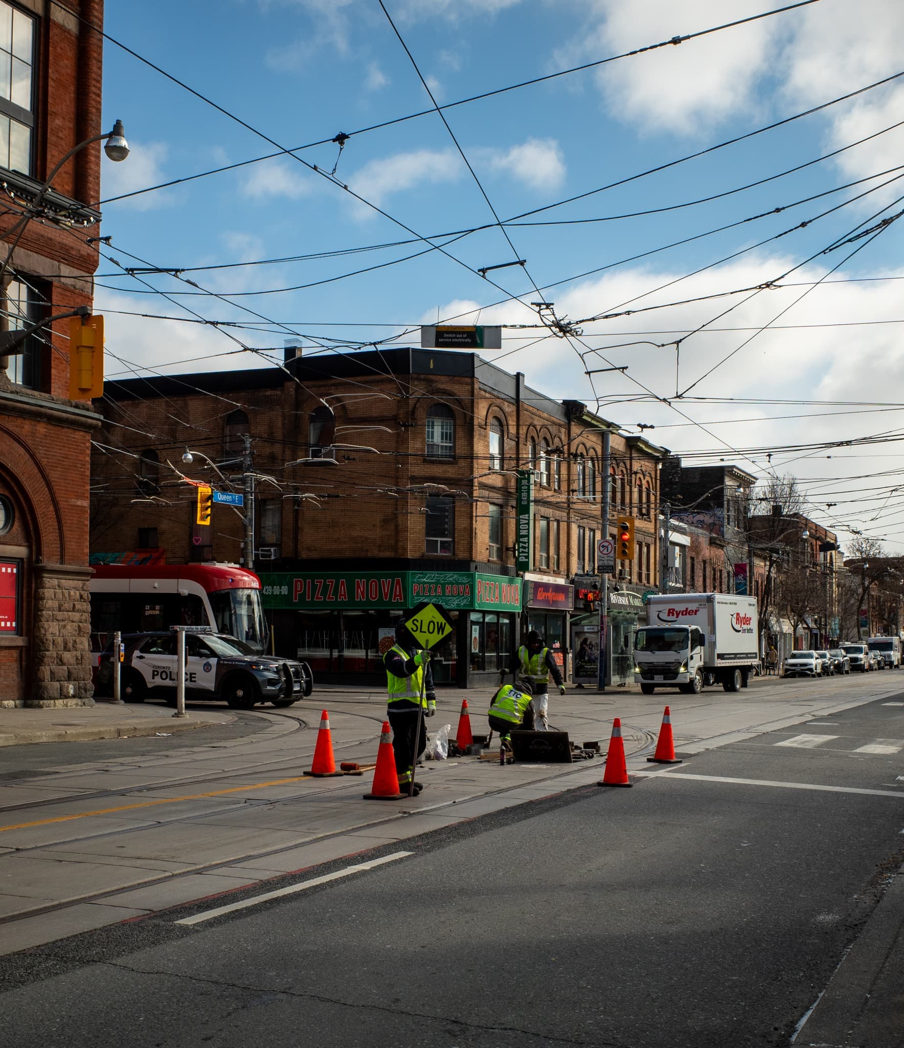 Workers in hi-vis vests kneel among traffic cones at a streetcar-track intersection, a red-brick storefront and truck behind