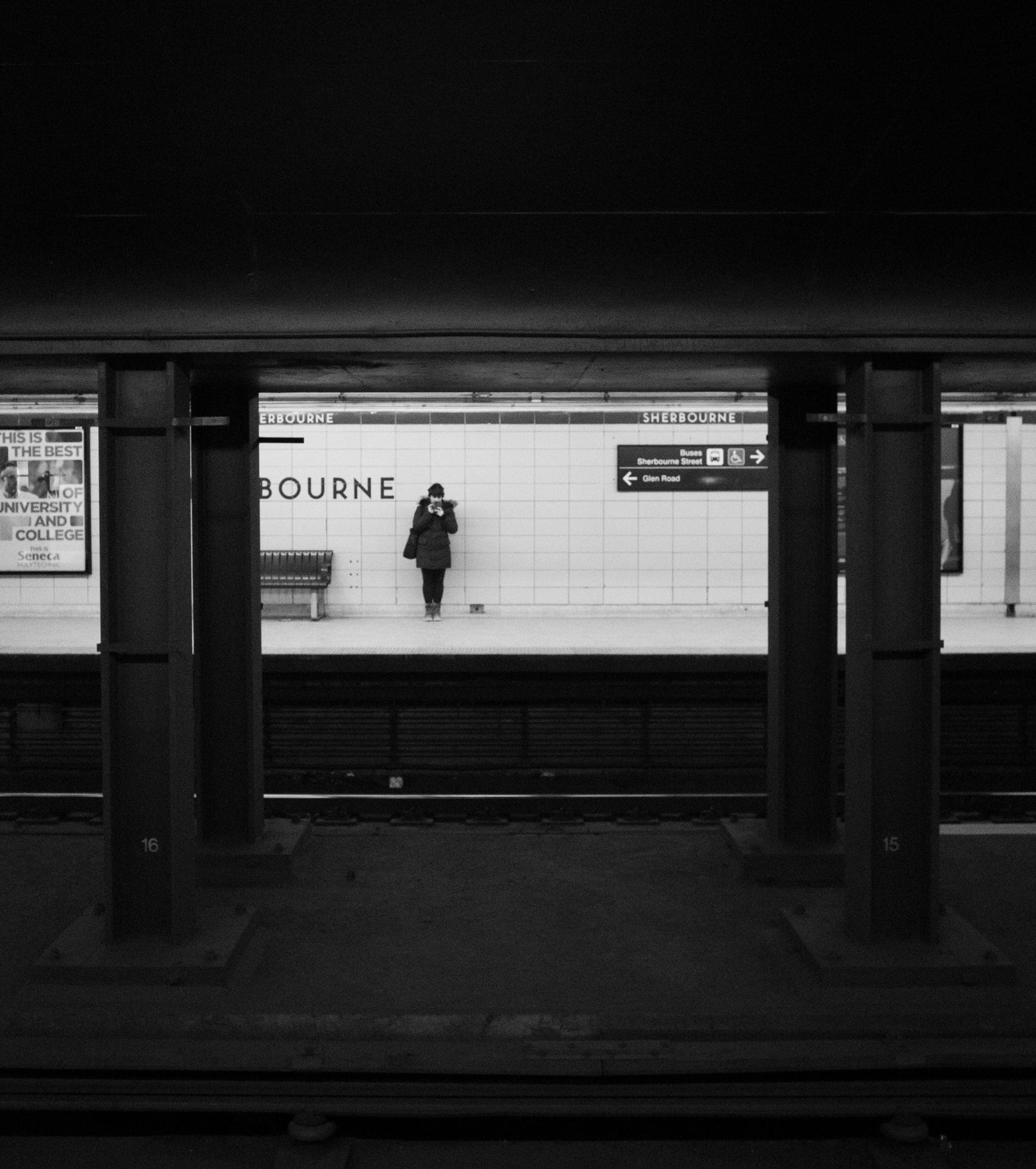 A lone figure in a dark coat at a tiled subway station, seen across the empty tracks, in black and white