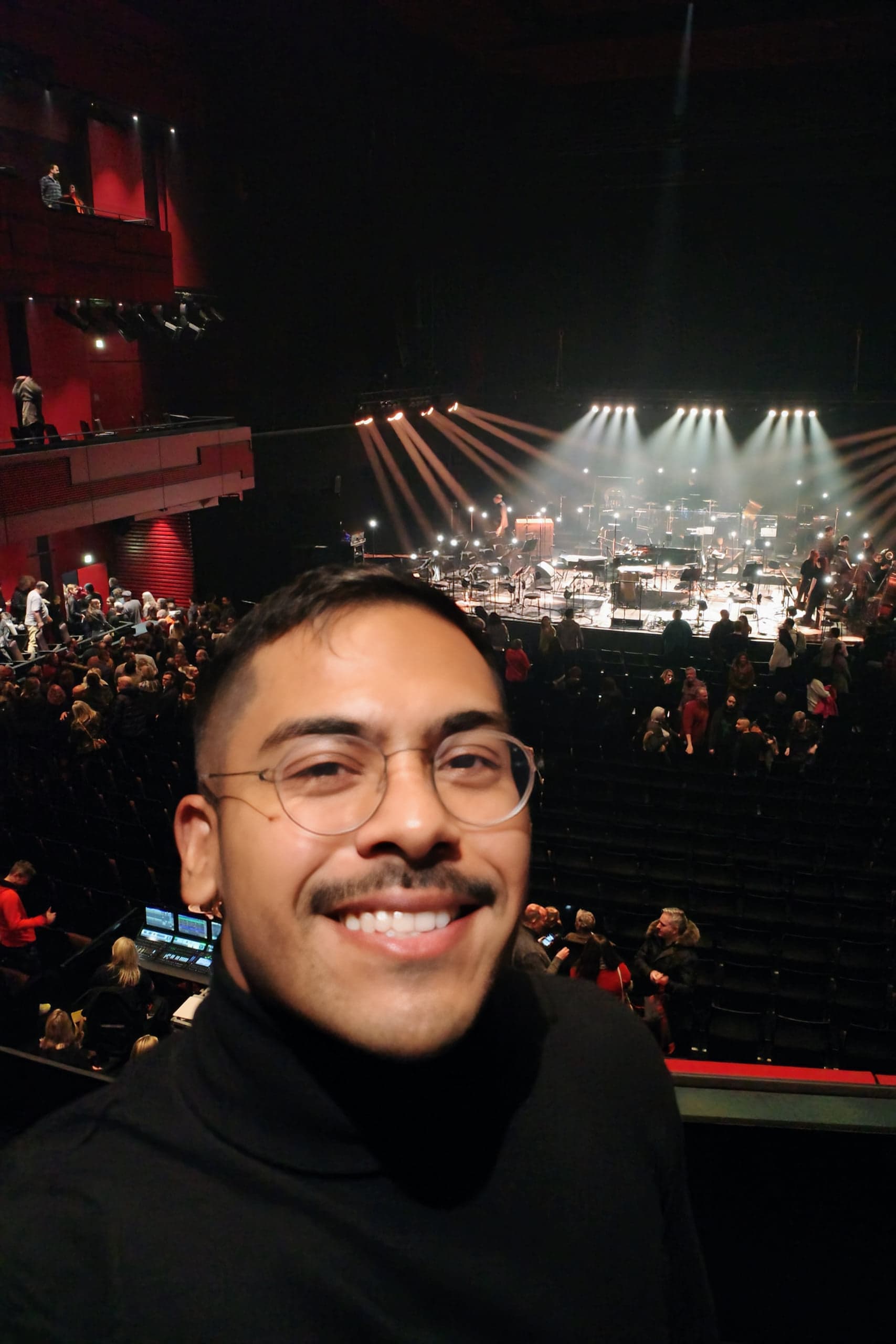A man in a black turtleneck and glasses smiles in a dark concert hall, stage and crowd behind him