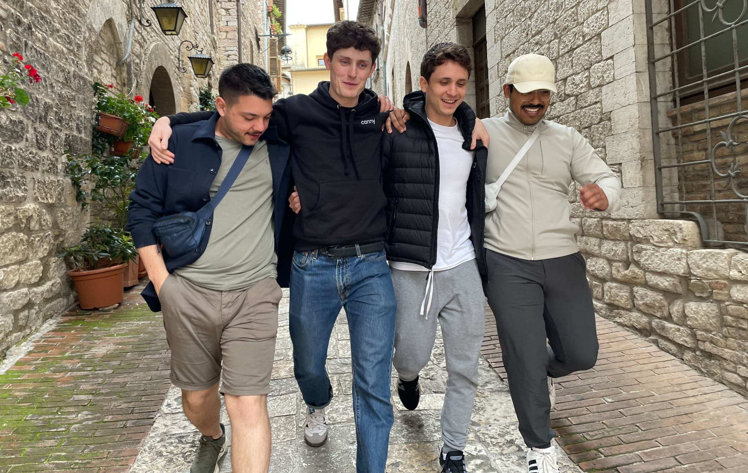 Four men walk arm-in-arm down a narrow cobbled street between old stone buildings