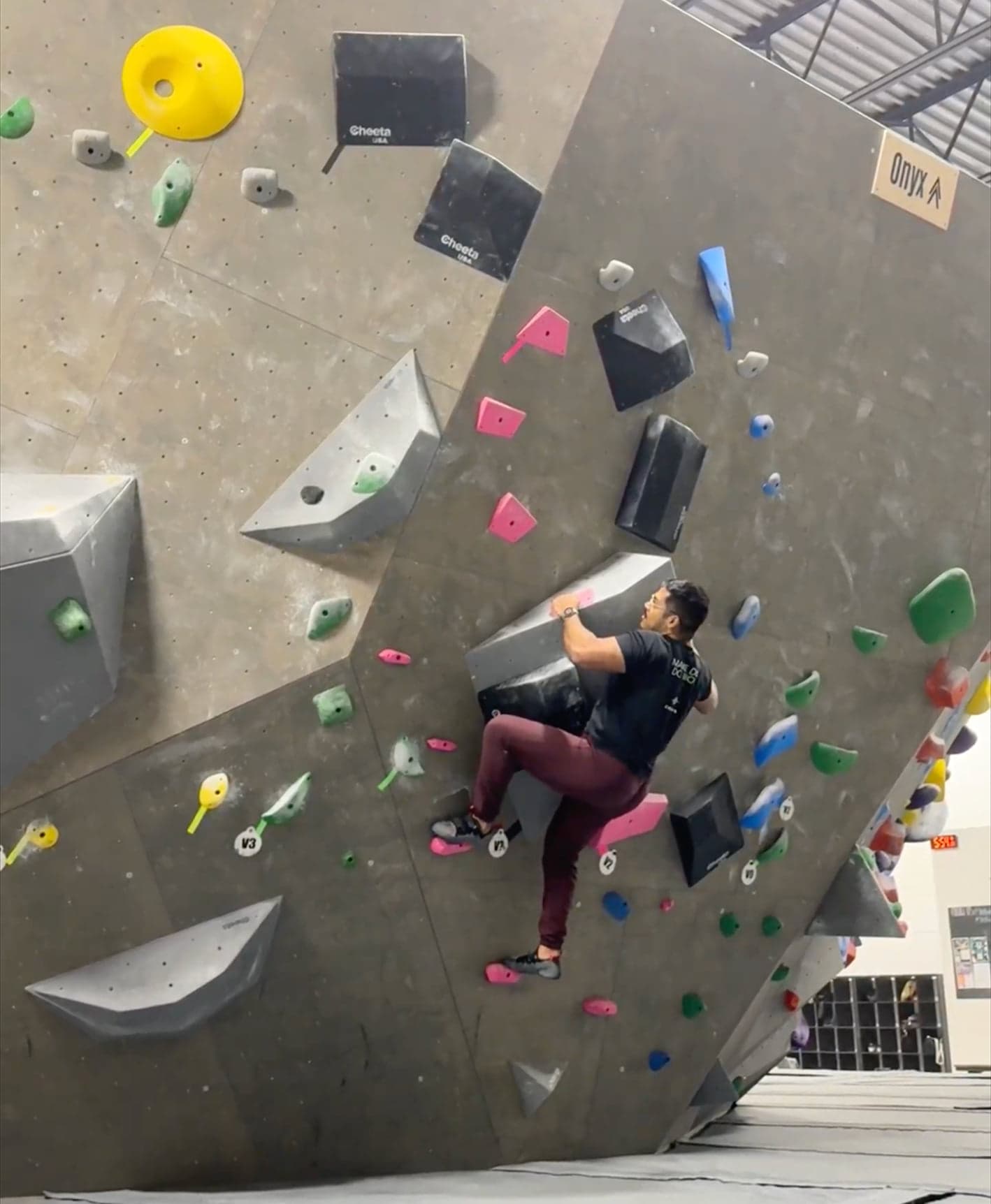 A climber in a black shirt and maroon pants grips holds on a steep indoor bouldering wall