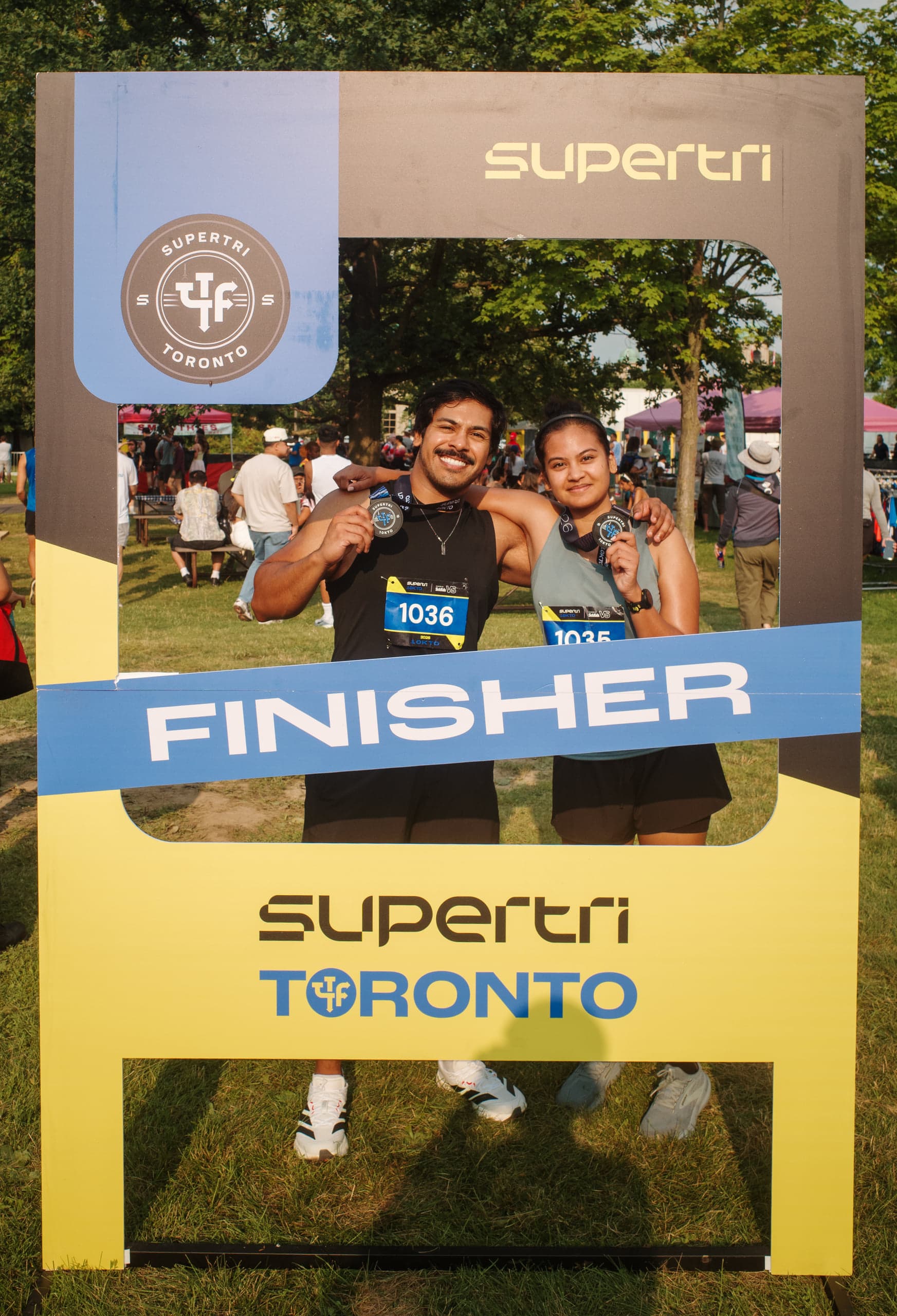 Two runners wearing race bibs hold up their finisher medals inside a large cutout finisher board on a grass field