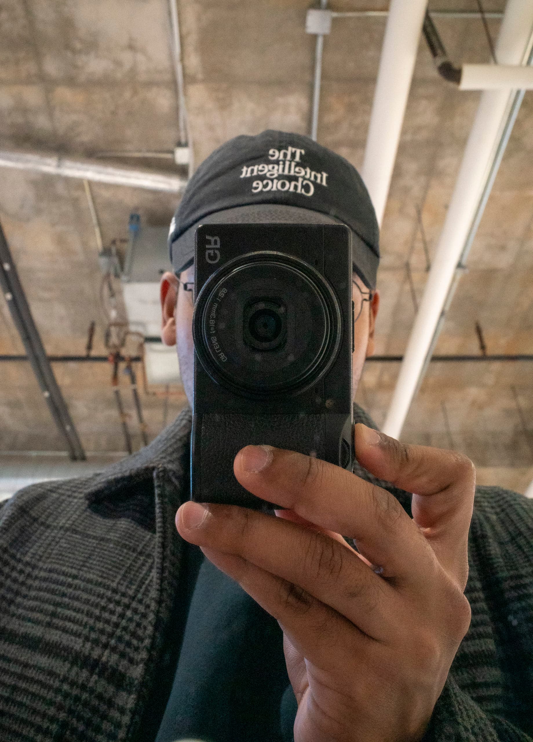 A person in a dark cap holds a small black compact camera to their face in a mirror reflection