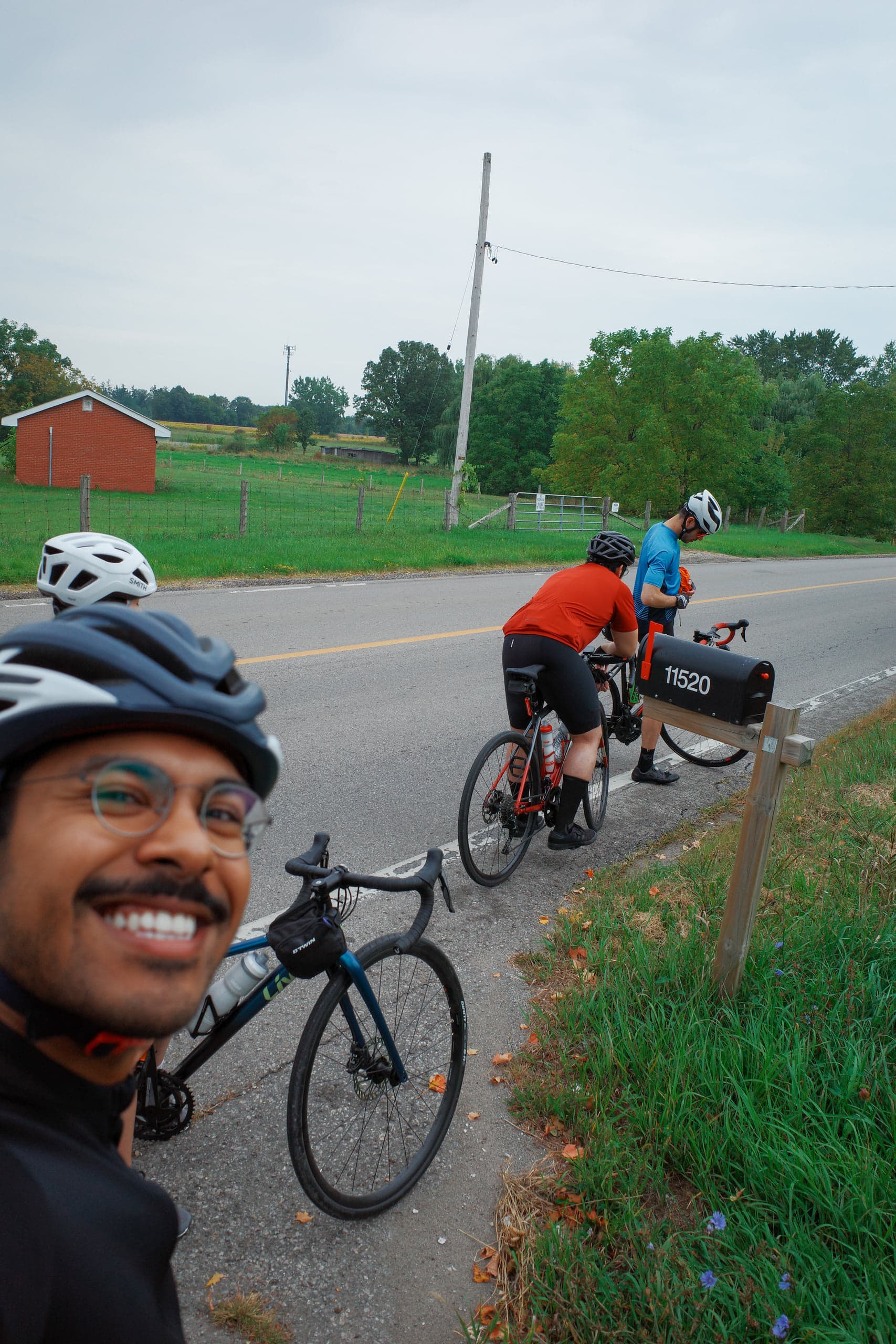 A cyclist in a helmet smiles in the foreground as two others stop by a roadside mailbox near farmland