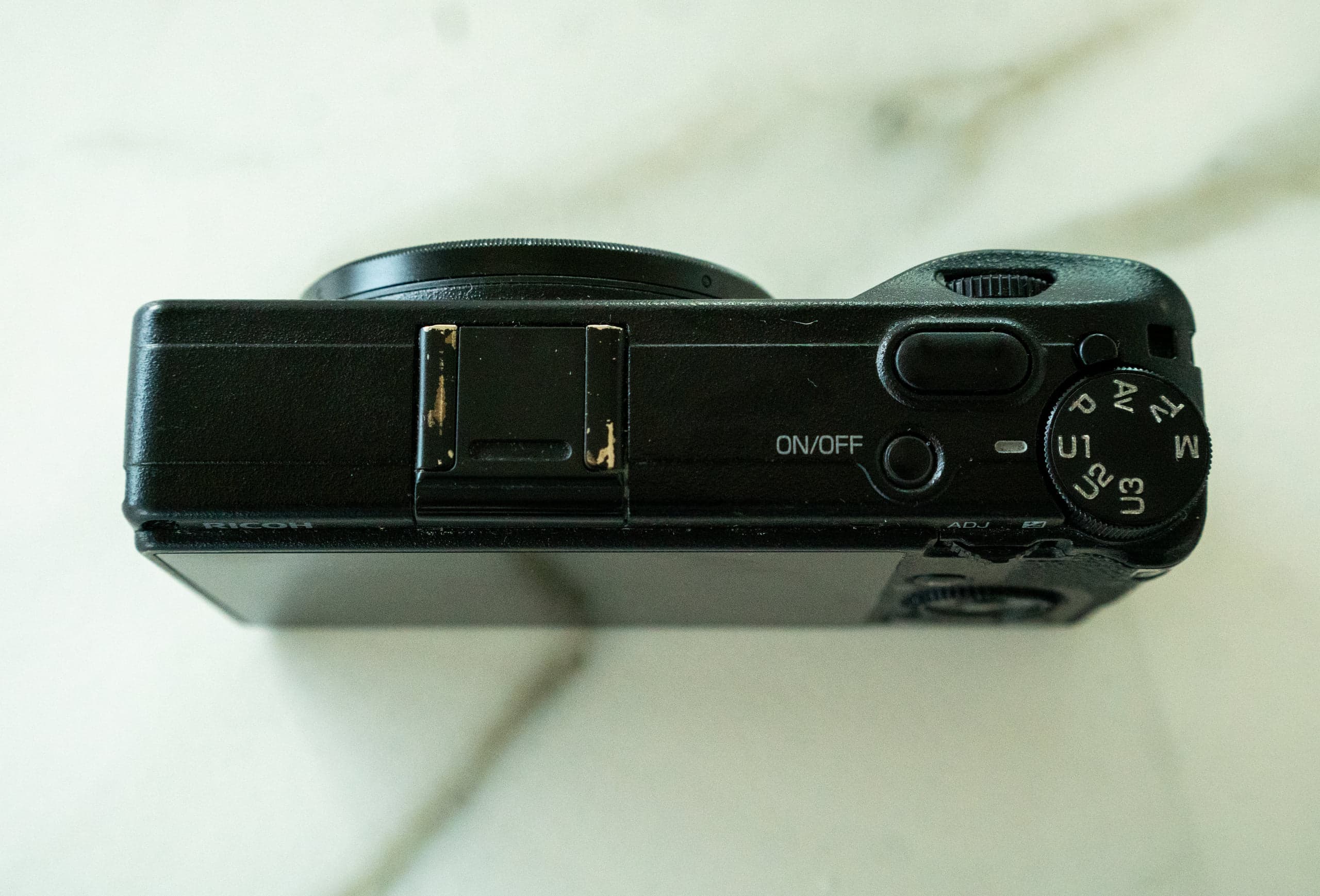 The top of a well-worn black Ricoh GR III on a marble surface, its mode dial turned to show U1, U2, U3, M, AV, TV and P, beside the ON/OFF button and a scuffed hot shoe.