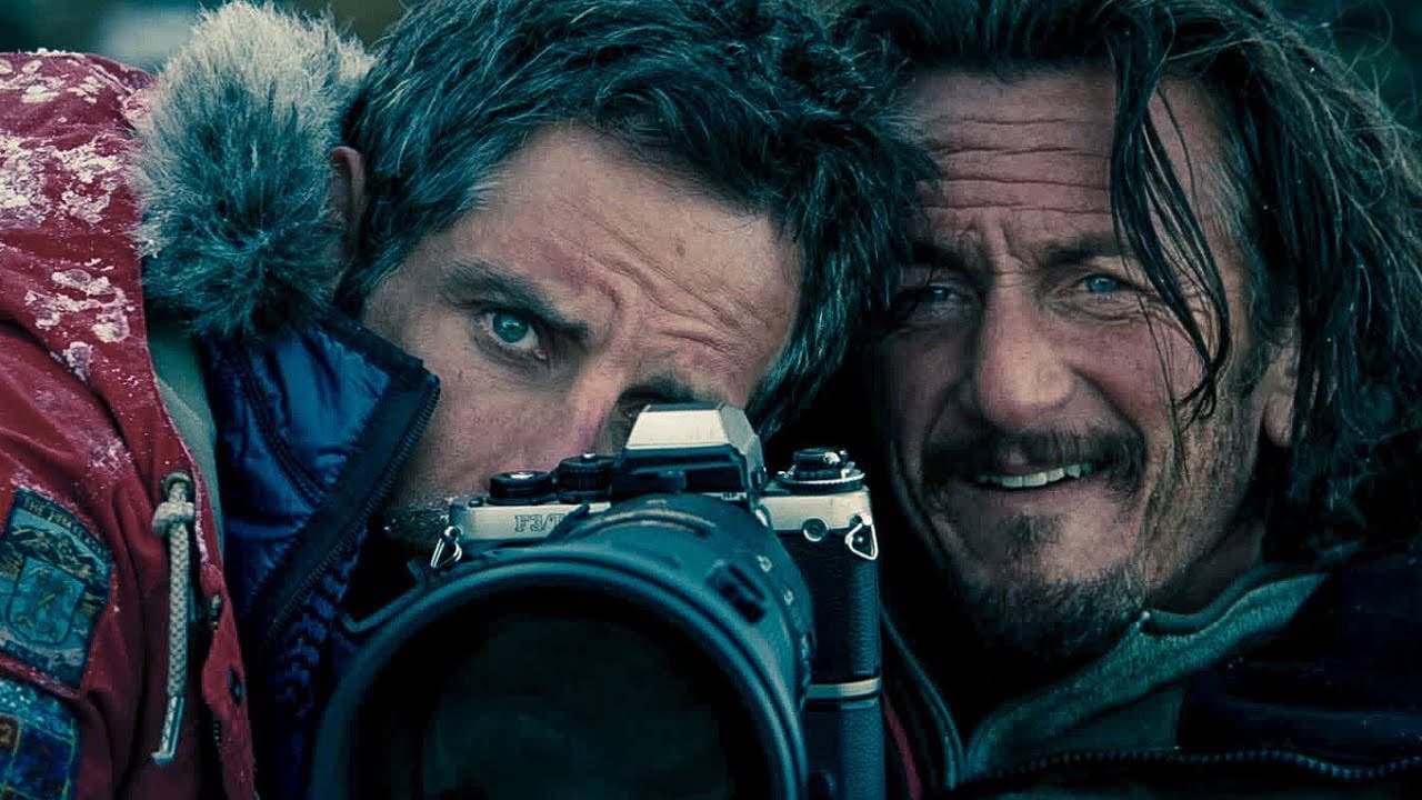 A still from The Secret Life of Walter Mitty: Walter Mitty, in a red fur-lined parka, peers through a film camera while Sean O'Connell leans in close beside him, both weathered by the cold on a snowy ridge.